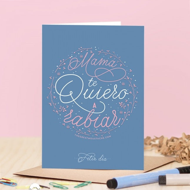 Tarjeta de felicitación azul con mensaje "Mamá te quiero a rabiar", sobre kraft, fondo de escritorio.